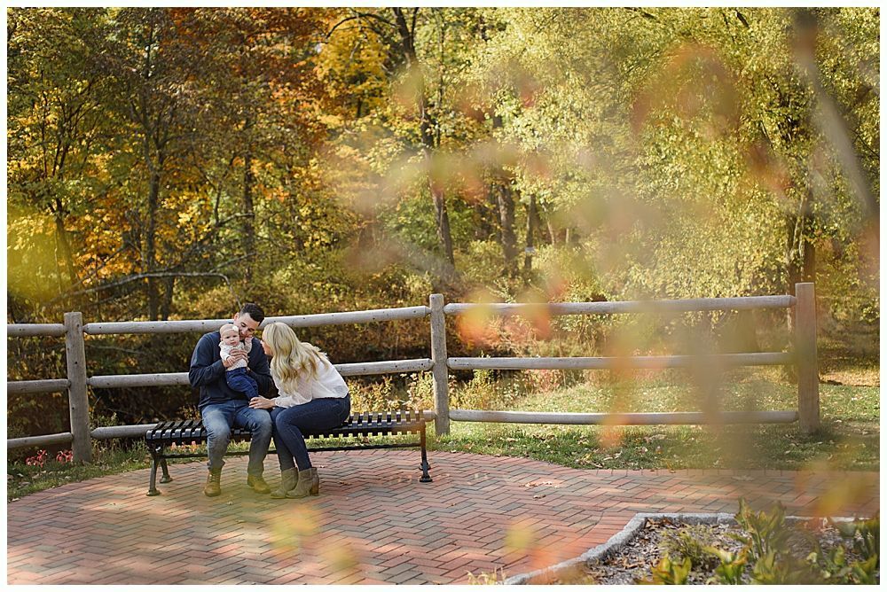 Father holding baby, both smiling. Blonde woman in foreground takes photo outdoors.