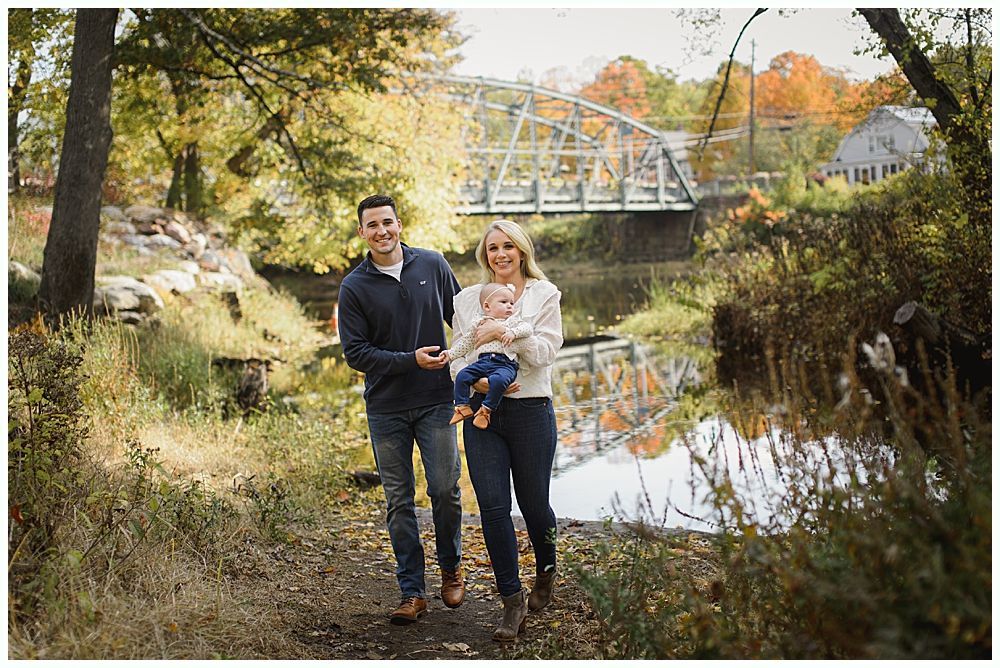 Family of three smiling while walking; bridge and fall foliage in the background.