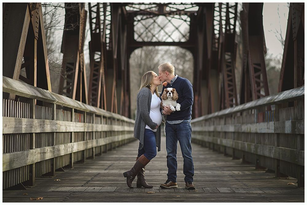 Couple kissing on a wooden bridge, holding a dog. Steel bridge in the background, fall colors.
