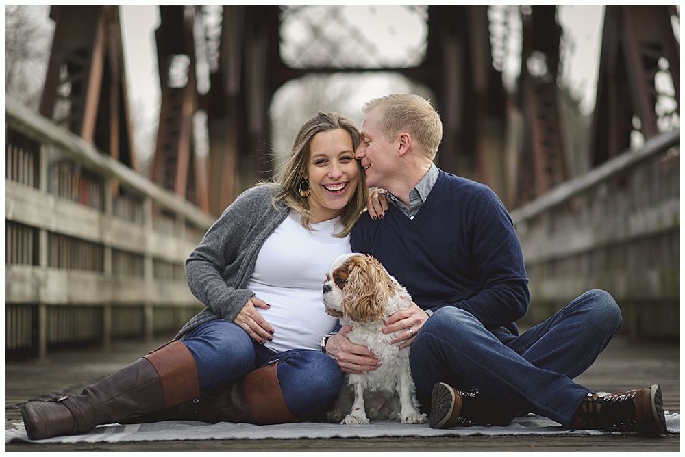 Pregnant woman and man seated with dog on bridge; woman smiles, man whispers.