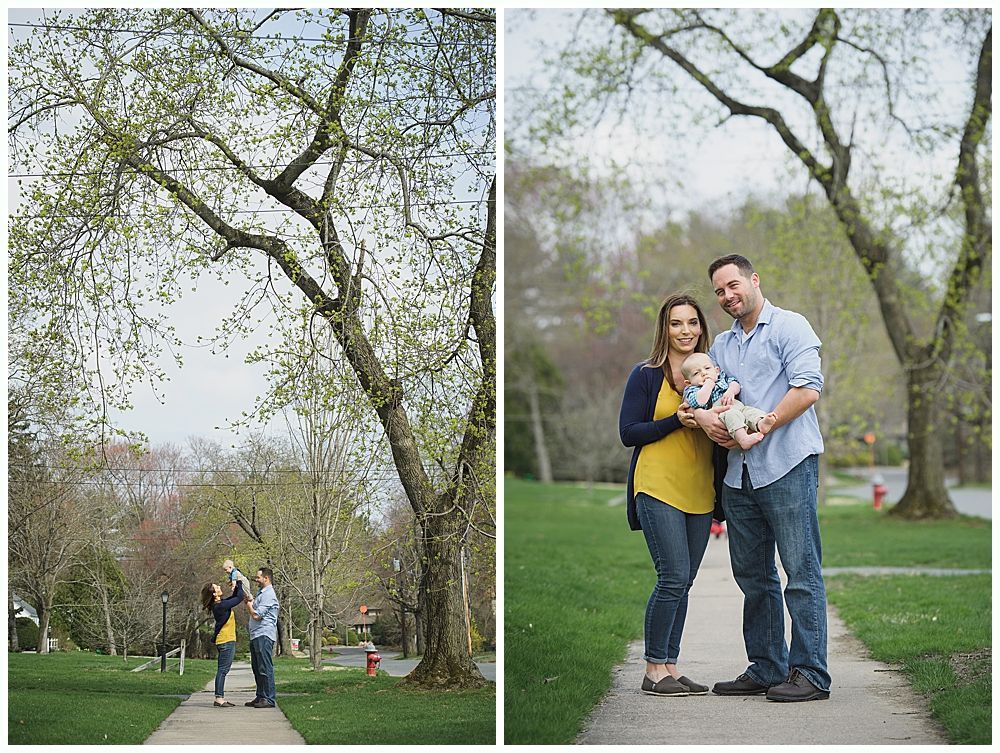 Family poses on a sidewalk under a tree with new leaves. The parents hold a baby.