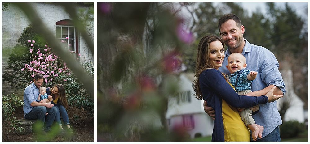 Family portrait, couple smiling, holding baby. Outdoor setting, house in background, green and pink foliage.