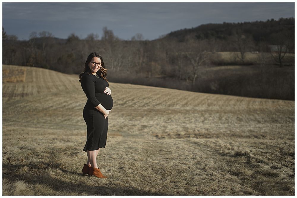 Pregnant person in black outfit stands smiling in a field with brown grass, trees, and hills.