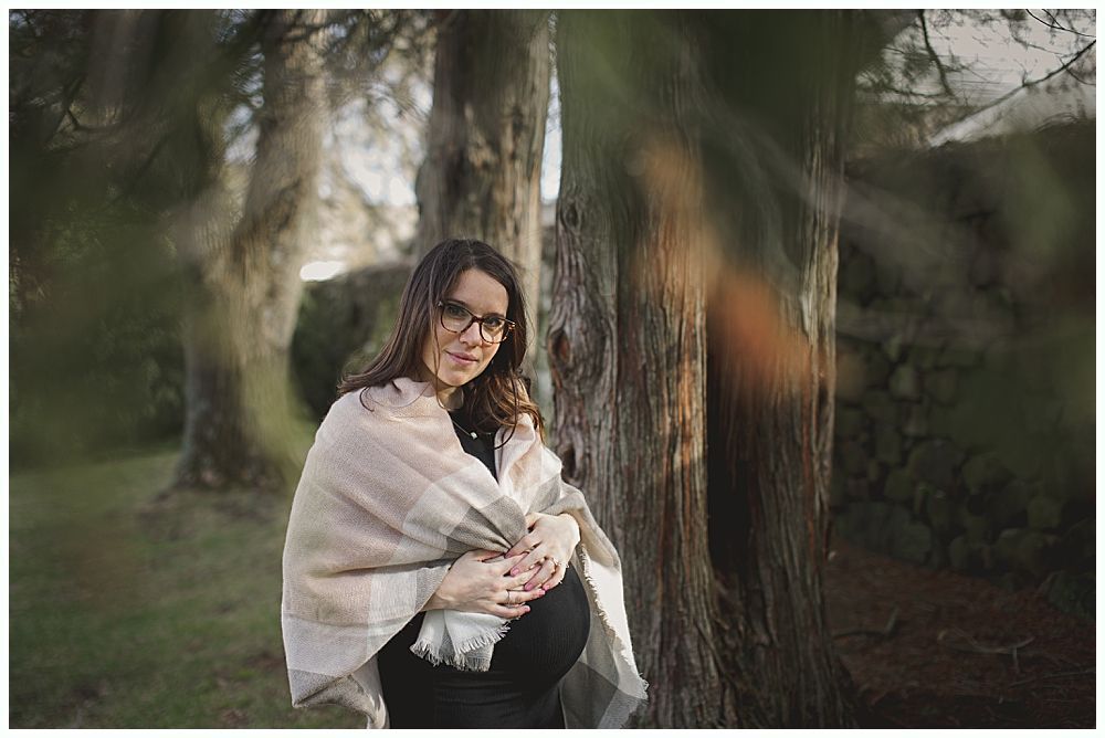 Pregnant woman in a field, holding her belly, wrapped in a blanket, looking at the camera. Trees in background.