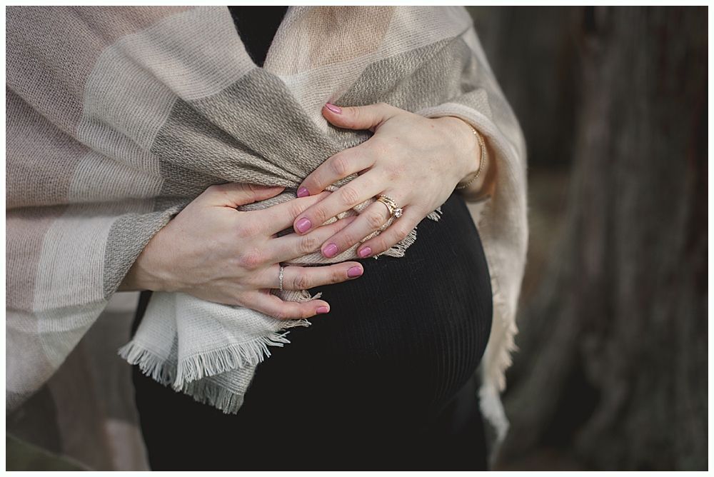 Pregnant person in black dress, hands cradling belly, wrapped in a plaid shawl.