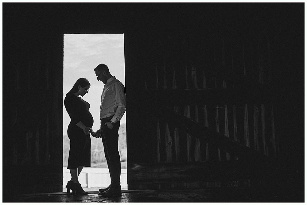 Pregnant person and partner silhouetted in a doorway, holding hands, in a dark barn-like structure.