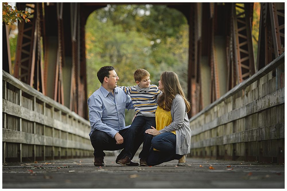 Family of three on bridge, smiling; fall colors, brown bridge, green background.