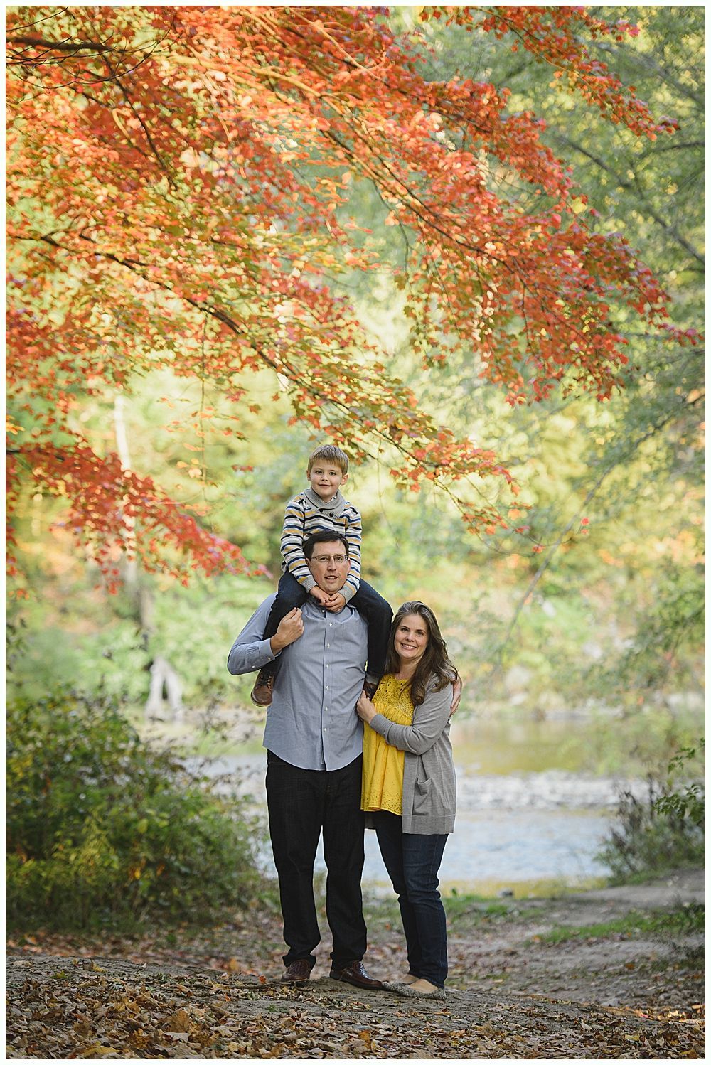 Family of three poses outdoors with red and orange foliage. Son on father’s shoulders, mother beside them.