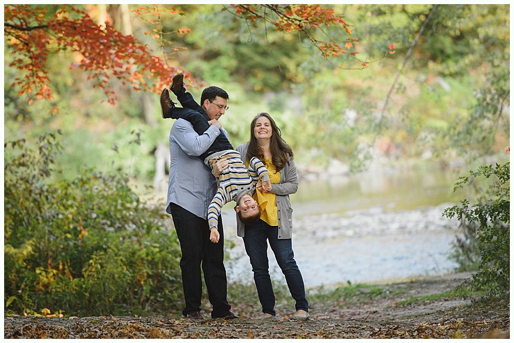 Family in fall setting: Father holding child upside down, mother smiling nearby.