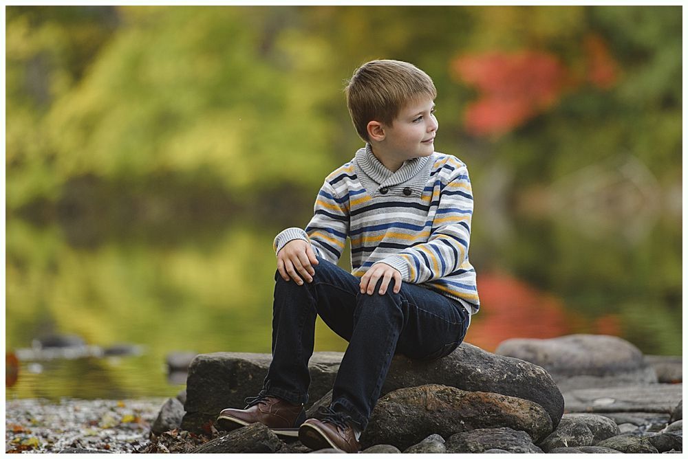 Boy sitting on a rock by water, looking to the side. Wearing sweater and jeans, autumn backdrop.