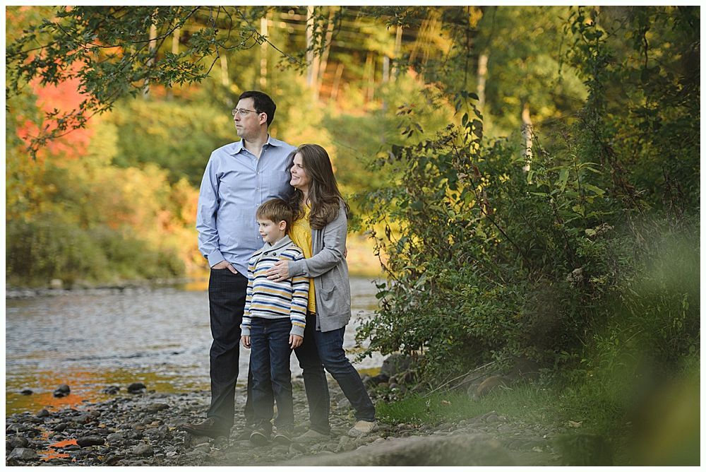Family standing near a river, fall foliage in the background. Man looking off, woman and child beside him.