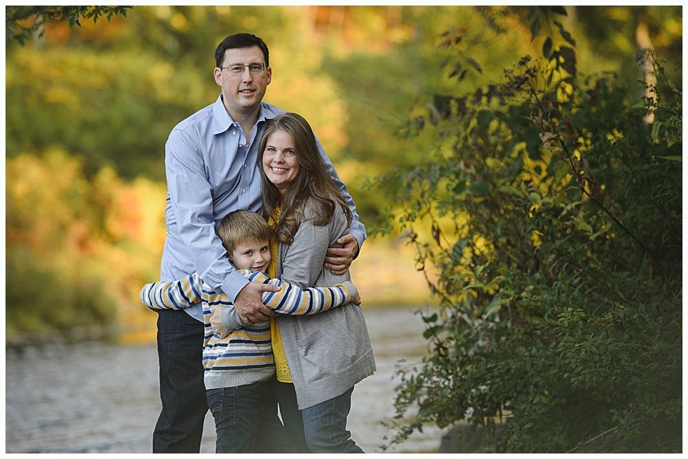 Family of three hugging outdoors near a river, fall foliage in background.