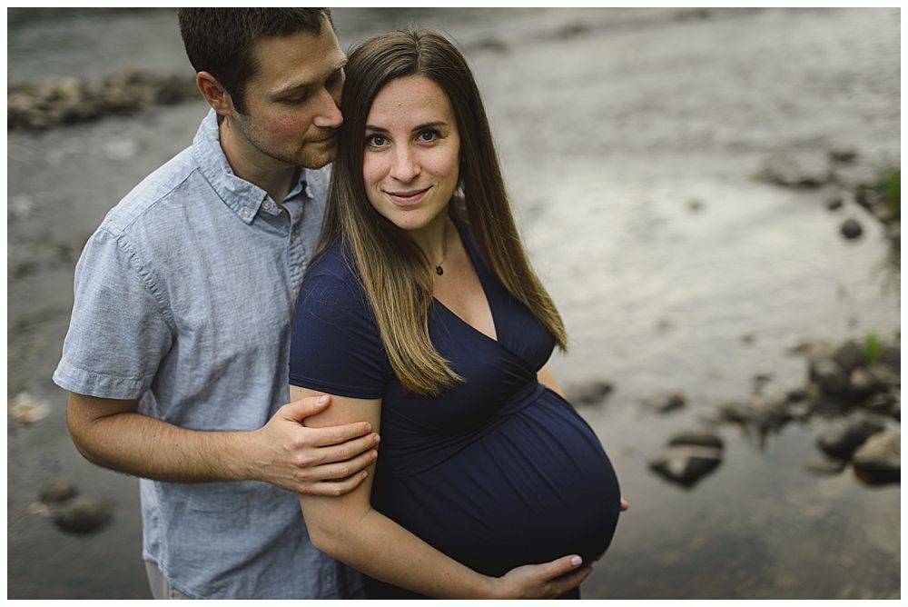 Couple poses outdoors; pregnant woman in blue dress, man in blue shirt, near a river.