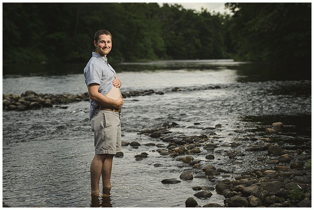 Man with a fake pregnant belly stands in a river, smiling, surrounded by trees.