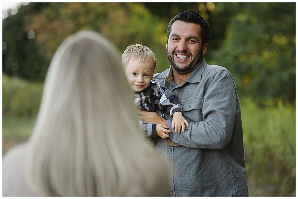 Father holding child, smiling, with mother blurred in the foreground. Outdoors with greenery in background.