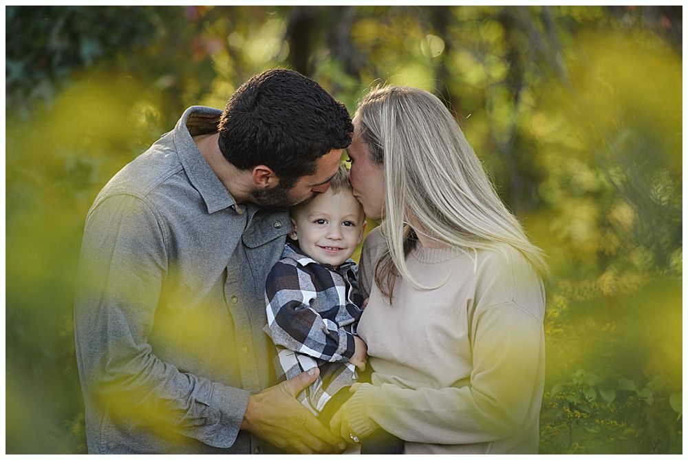 Parents kissing the heads of their smiling child. Outdoors, soft focus with green foliage.