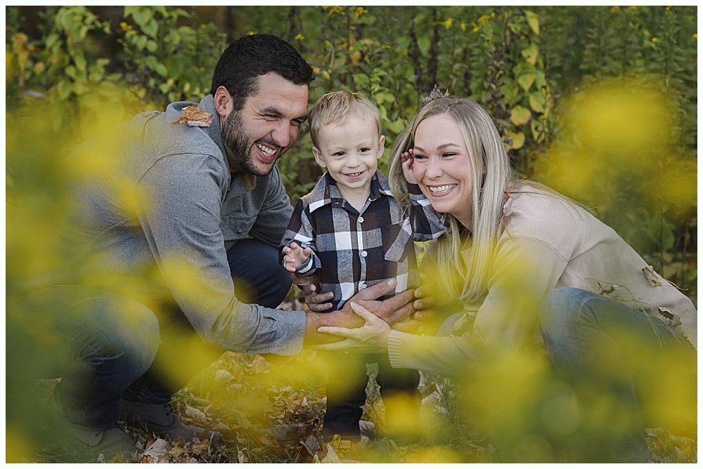 Parents kissing the heads of their smiling child. Outdoors, soft focus with green foliage.