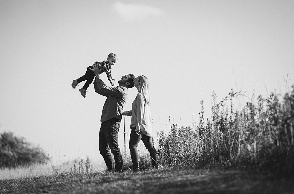Father lifting child, mother watches, sunny outdoor field.
