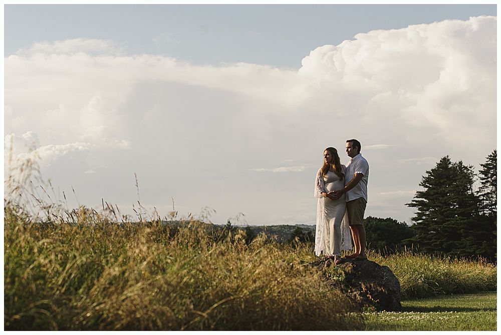 Couple embracing, expecting, in grassy field. Pregnant woman, man holding her, looking at sky with clouds.