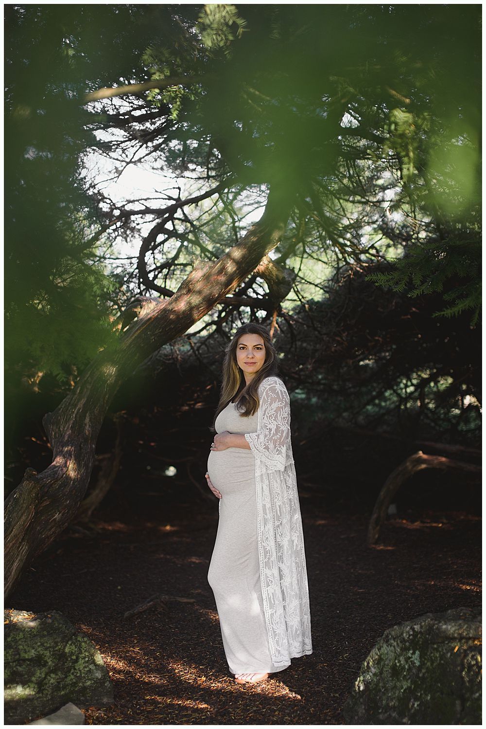 Pregnant woman poses in a forest, wearing a white dress and lace shawl, hand on her belly.