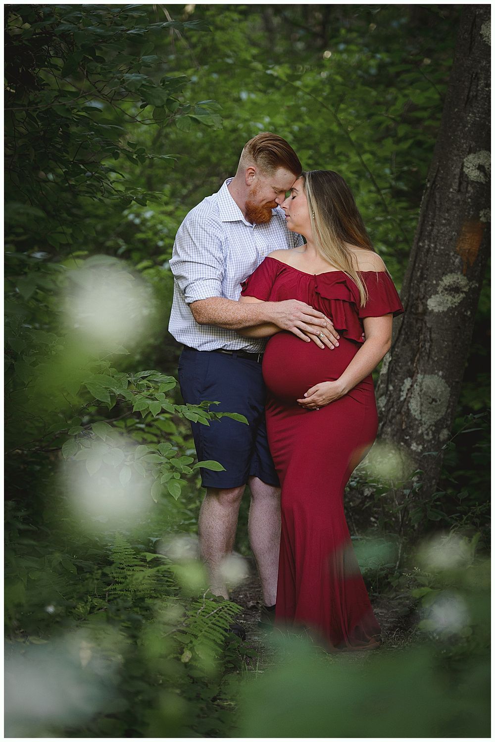 Pregnant woman in red dress with man, outdoors, forest setting, embracing.