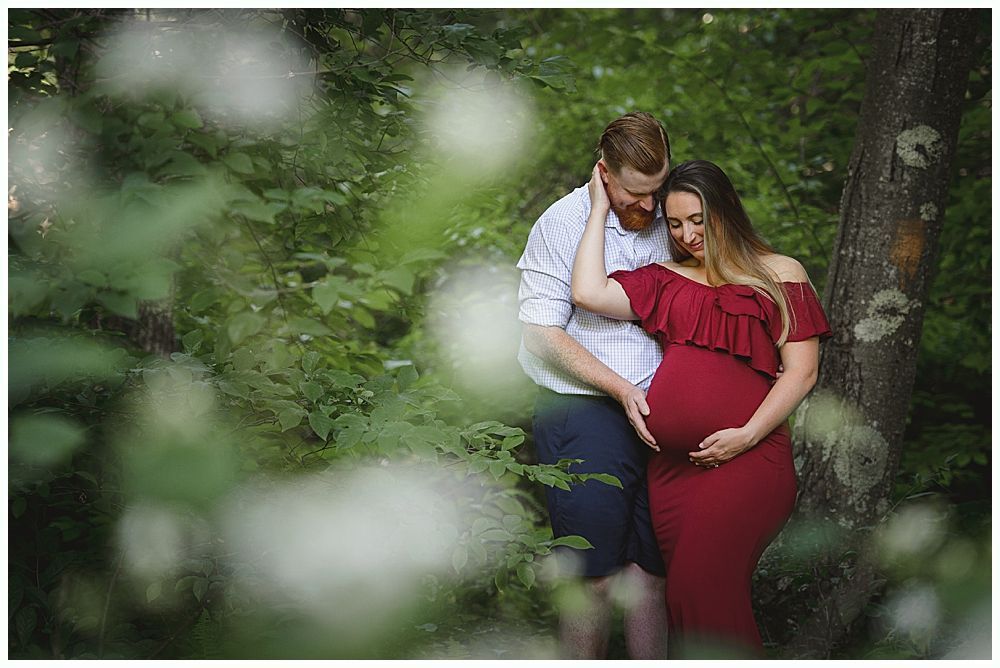 Couple in forest, woman cradling baby bump, man holding her arm; both look down.