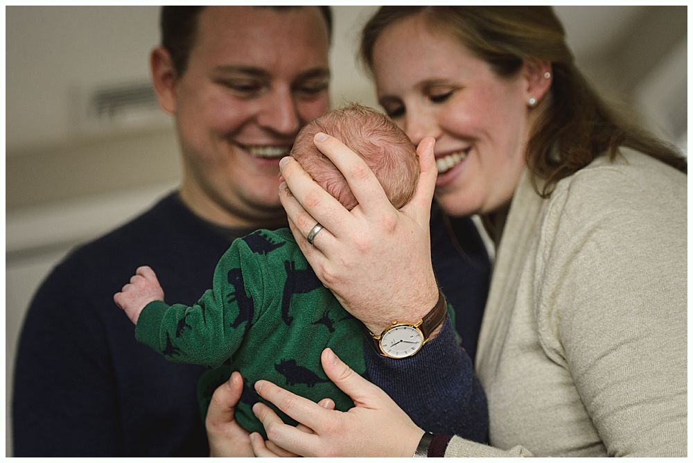 Parents smiling, holding a newborn baby wearing a green patterned onesie.
