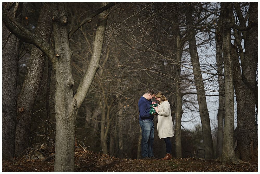Couple embracing in a forest with bare trees, wearing blue shirt and beige coat.