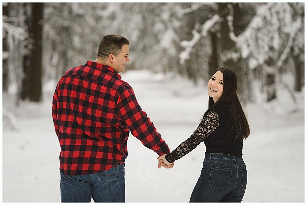 Couple holding hands, walking in a snowy forest; man in red plaid shirt, woman smiling with black top.