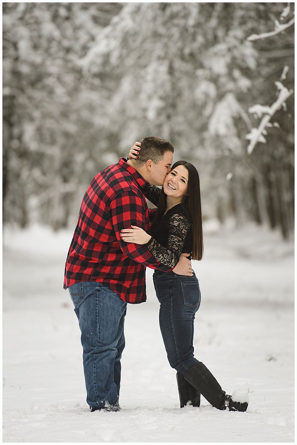 Man kissing woman's cheek in snowy forest. Both are smiling, he wears a red plaid shirt, she a black top.