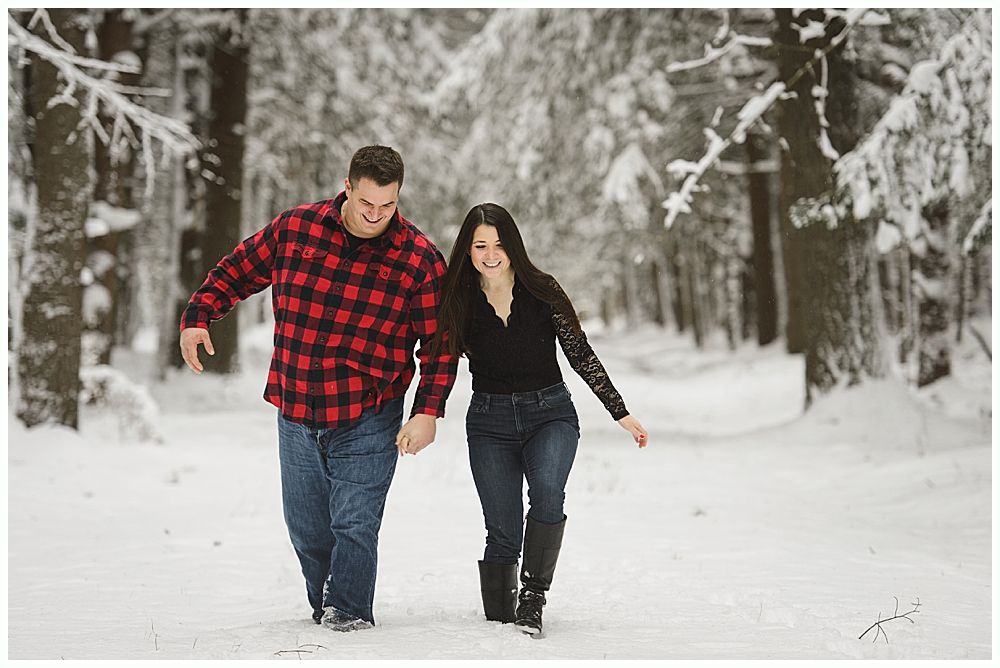 Man kissing woman's cheek in snowy forest. Both are smiling, he wears a red plaid shirt, she a black top.
