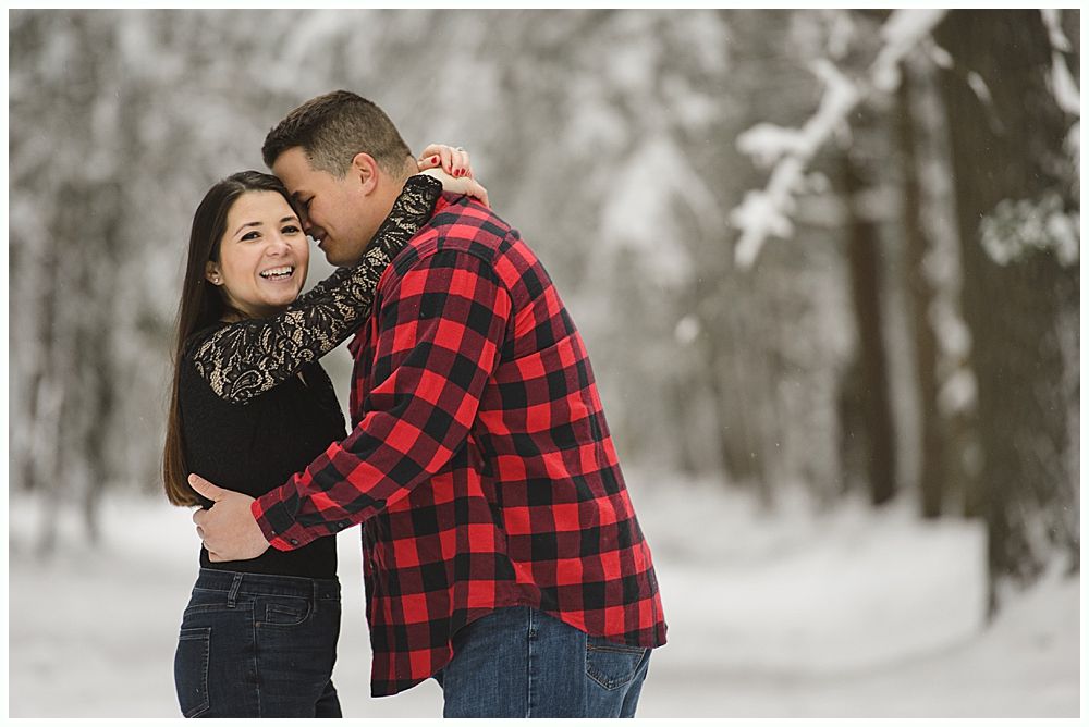 Couple embracing in snowy woods; man in red plaid shirt, woman in black top smiles.
