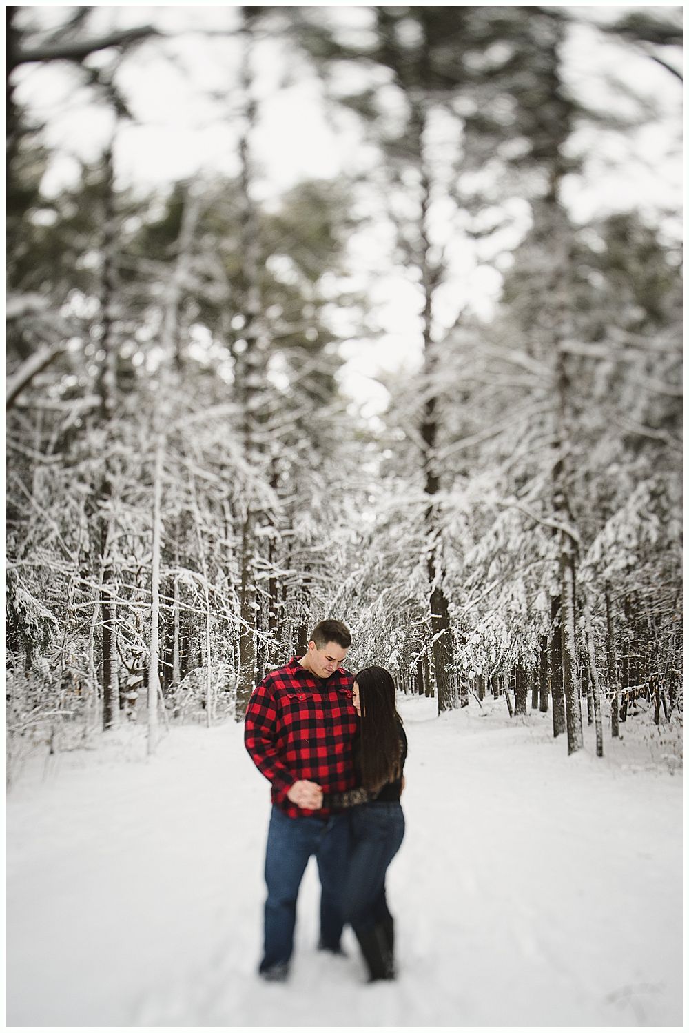 Couple embraces on a snow-covered path, surrounded by snow-laden trees. Man in red plaid shirt, woman in black coat.