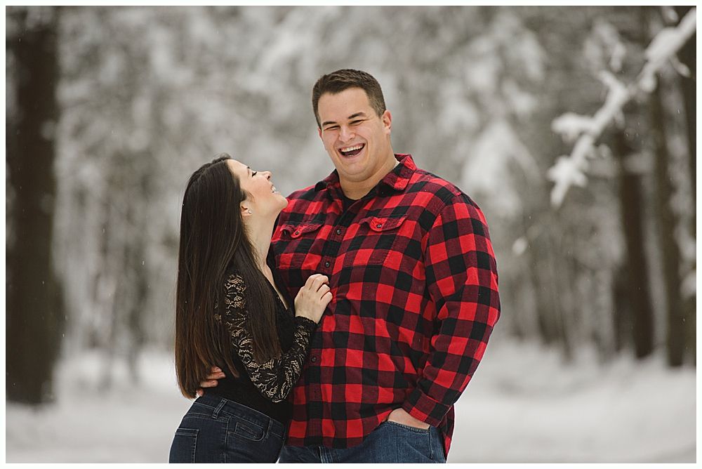 Couple laughs in a snowy forest. Woman in black lace top, man in red plaid shirt.