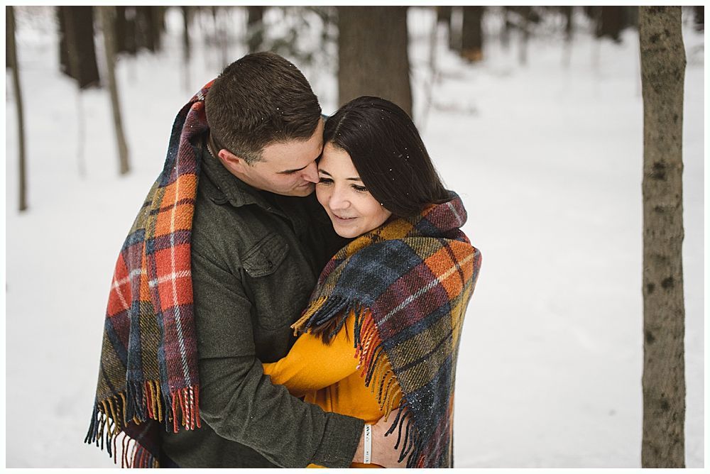 Couple embraces in a snowy forest, wrapped in a plaid blanket. Man kisses woman's cheek; both are smiling.