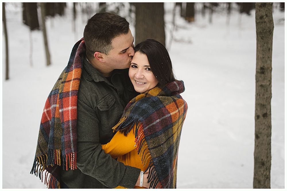 Couple embracing in a snowy forest, man kissing woman's forehead, wrapped in a plaid blanket.
