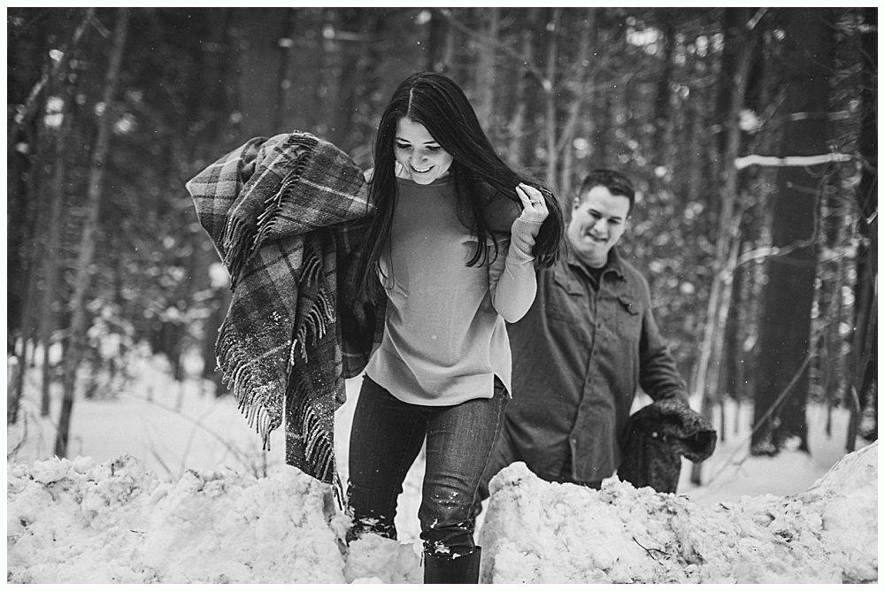 Woman carrying blanket, walking in snow; man follows behind in wooded area. Black and white photo.