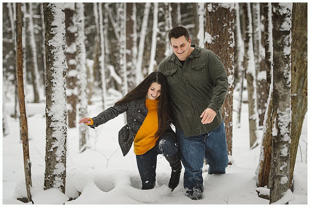 Couple walking through snowy woods, man smiling, woman looking down, both in winter attire.