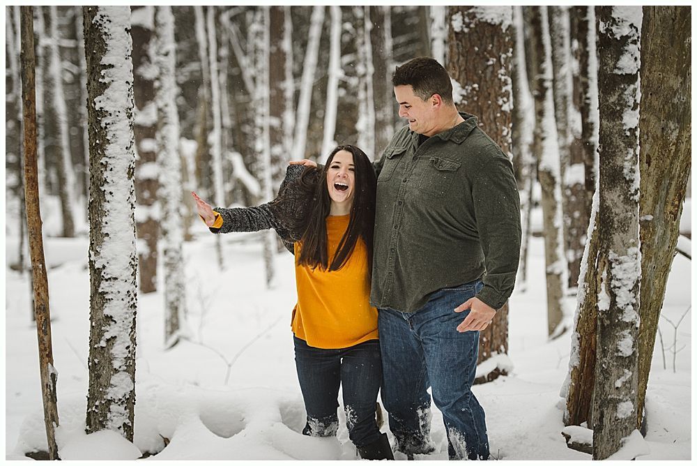 Couple in snowy woods; woman with open mouth and arms out, man looks on, trees.
