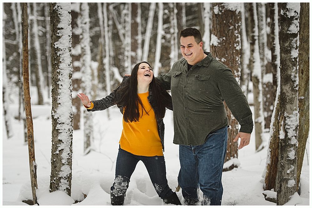 Couple laughs in snowy forest, woman with arms raised, man with arm around her shoulders.