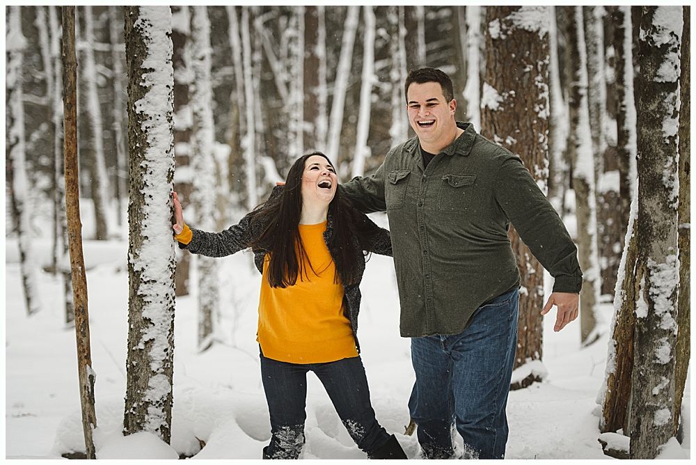 Couple laughing in a snowy forest. Woman in yellow, man in green, embracing near trees.