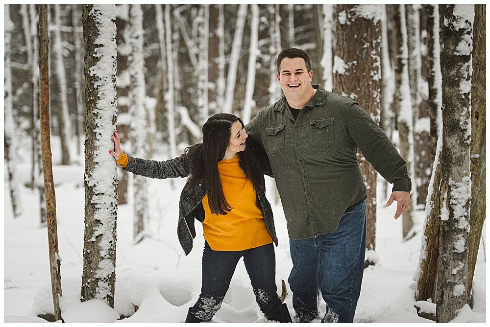 Couple smiling, posing in snow-covered forest. Man in green shirt, woman in yellow shirt, trees surround them.