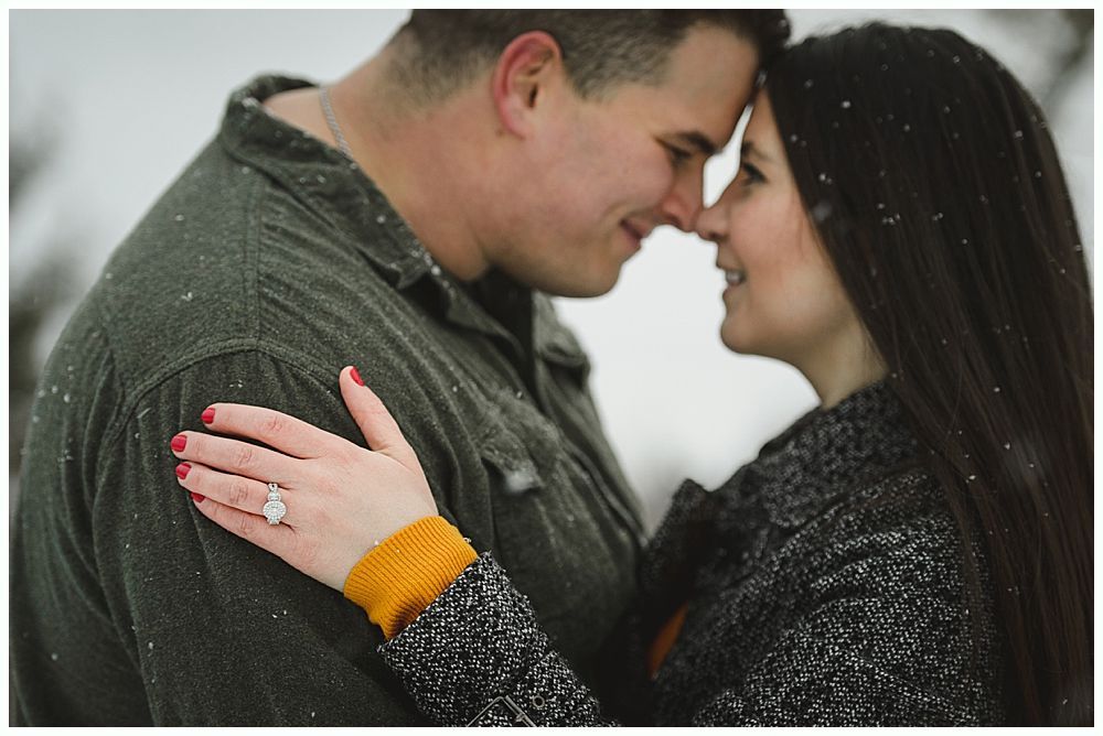 Couple embracing, noses touching, looking at each other. Snowy outdoor setting. Woman's hand with engagement ring visible.