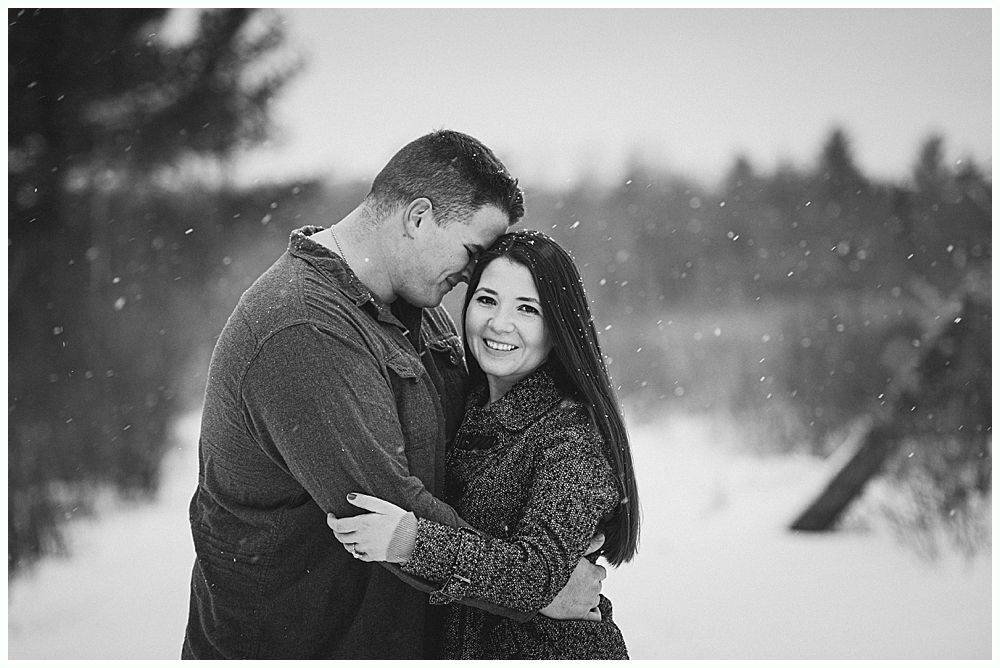 Couple embraces in snowy field; woman smiles, man in background. Black and white.