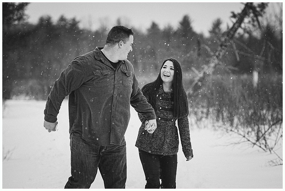 Couple walking hand-in-hand in snowy woods, smiling. Snow falling.