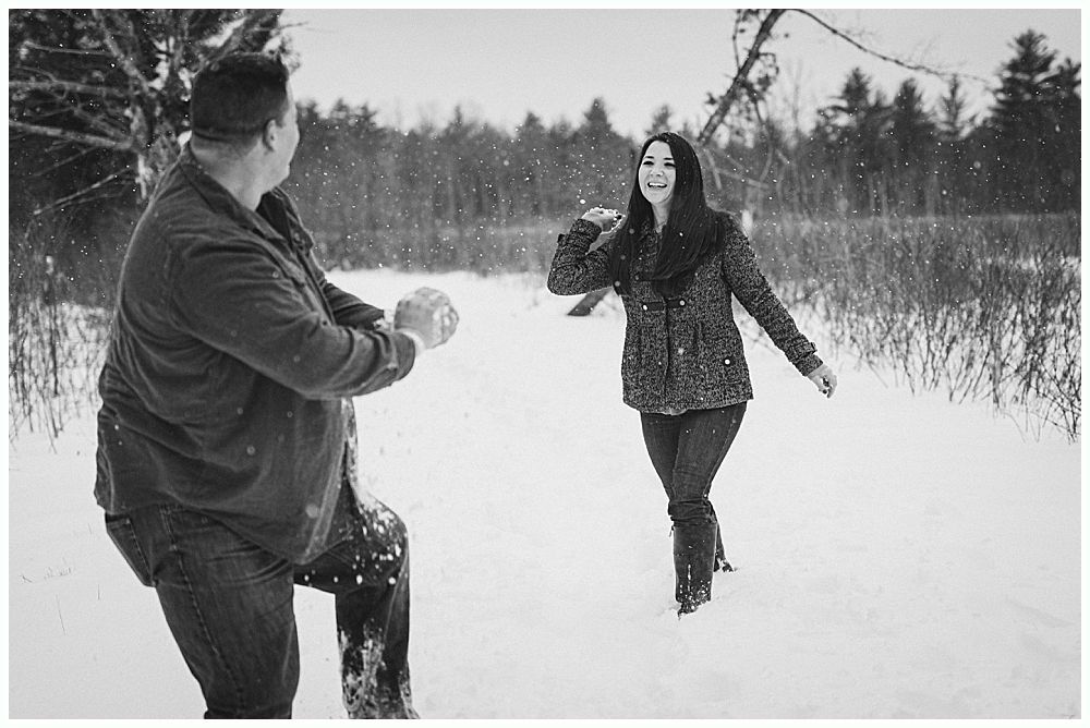 Man throws snowball at woman in a snowy forest; she smiles and reaches her arm up.