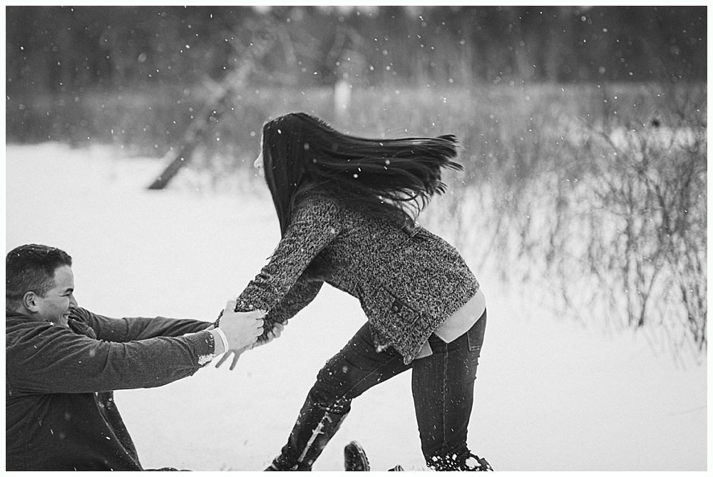 Couple playfully pulling on each other's hands in snowy landscape; one with long hair waving.