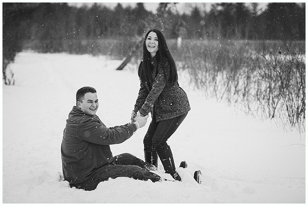 Couple playing in the snow; woman helps man up as they laugh outdoors on a snowy day.