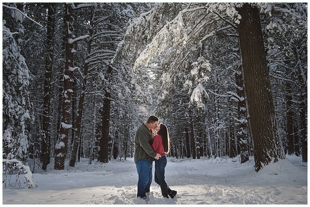 Couple embraces in snowy forest, sunlight streams through trees.