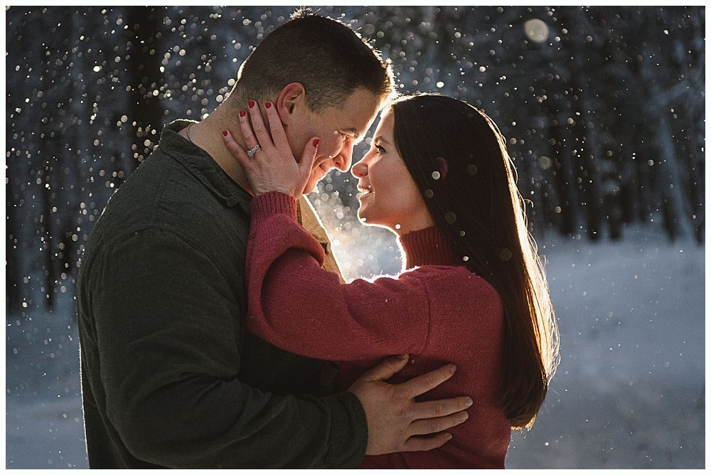 Couple embraces in a snowy landscape. Woman touches man's face, both smiling. Sunlight glows behind them.